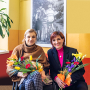 Žena sedící na vozíku s kyticí v ruce a vedle ní dřepící žena s květinami, obě se usmívají, v pozadí visí na zdi černobílá fotografie cyklistů.