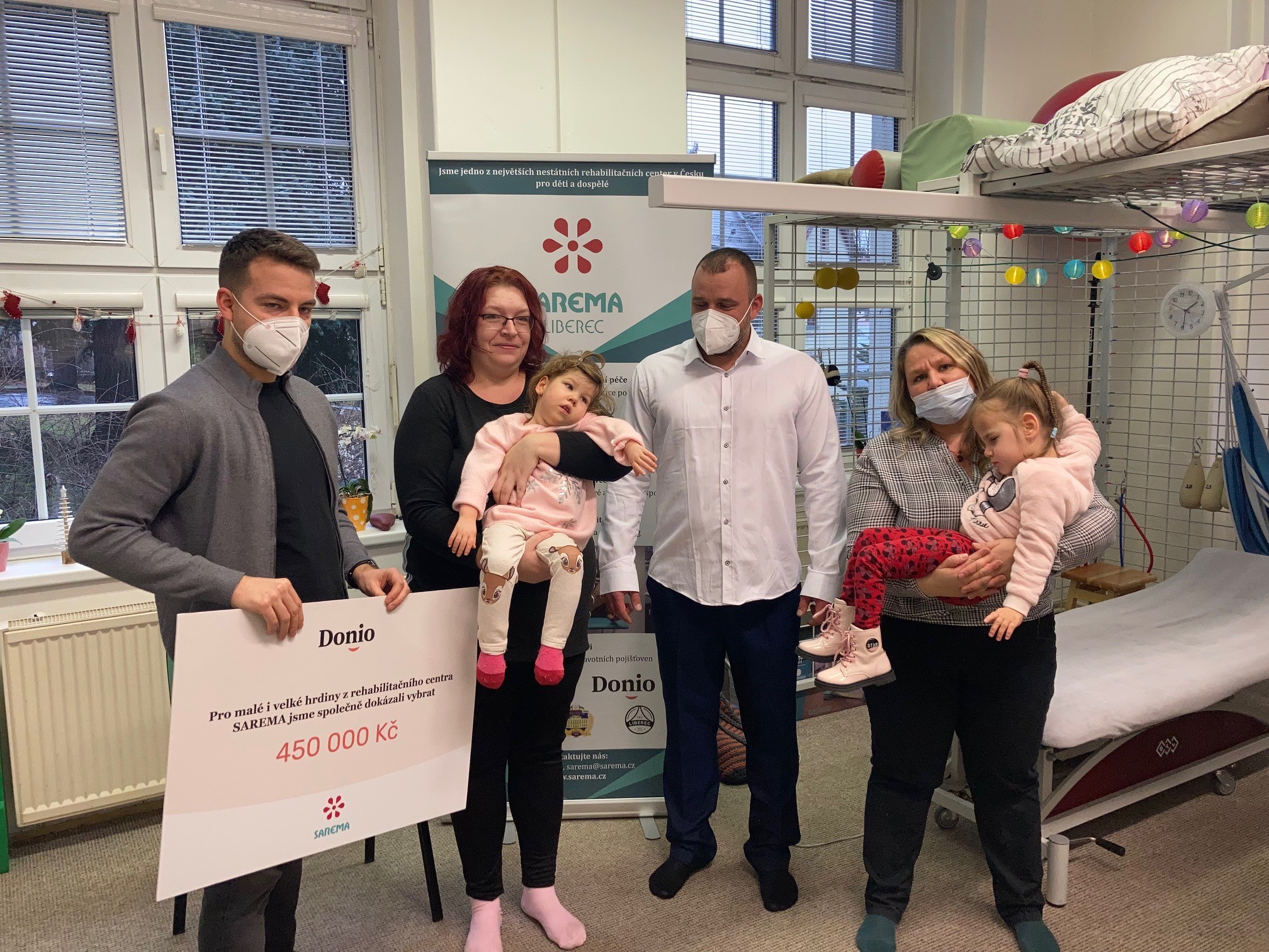 Skupina dospělých s dvěma malými dětmi v náručí, focená uvnitř rehabilitačního centra SAREMA Liberec, vedle banneru a symbolického šeku