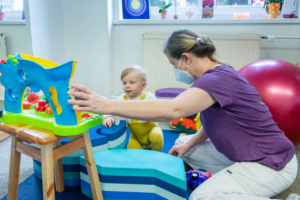 Terapeutka v kleku jistí batole u pěnových stupínků během cvičení s hračkou