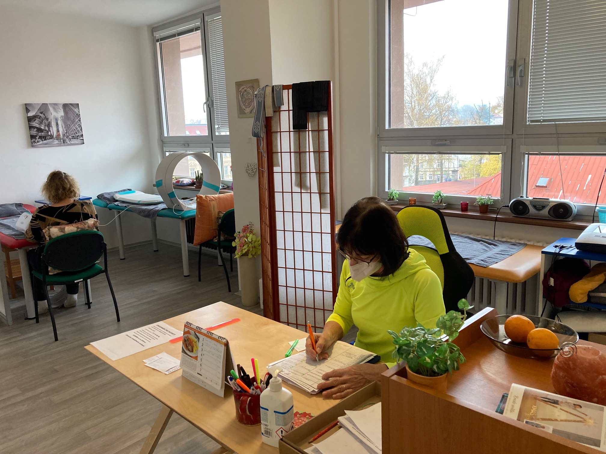 Recepce v rehabilitačním zařízení, žena s respirátorem zapisuje do diáře, v pozadí pacientka na lehátku