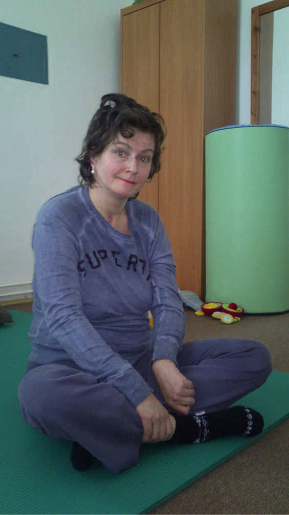 Žena sedící na podložce v místnosti pro rehabilitaci, oblečená v teplákové soupravě s nápisem Superdry