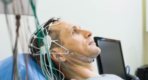 Detail hlavy muže vleže s elektrody připevněnými k lebce a bradě při neurologickém vyšetření, v pozadí monitor.