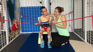 Chlapec v rehabilitačním obleku v kleci Spider s fyzioterapeutkou, která ho podporuje a usmívá se na něj.