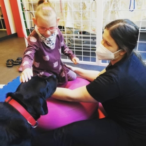 Dívka při rehabilitaci na míči se usmívá, hladí černého psa při canisterapii a je jištěná terapeutkou v respirátoru.