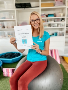 Usměvavá žena v tyrkysovém tričku SAREMA sedí na gymnastickém míči a drží certifikát ochranné známky