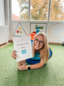 Žena v tričku Sarema leží na zeleném koberci a s úsměvem drží certifikát ochranné známky.