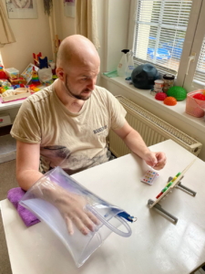 Muž procvičuje motoriku ruky pomocí speciální rehabilitační pomůcky s průhlednou komorou a barevnými prvky.