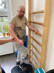 Muž cvičí rovnováhu na rehabilitační pomůcce u žebřin s dopomocí terapeuta.