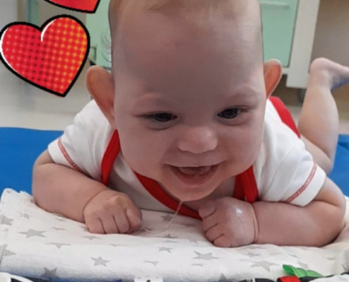 Miminko s vývojovým postižením leží na břiše a usmívá se, vedle něj jsou dvě kreslená srdce.